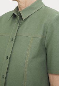 Camicia verde a maniche corte con colletto, due bottoni di chiusura e cuciture contrastanti in tan lungo le sue cuciture. Tessuto morbido e textured.