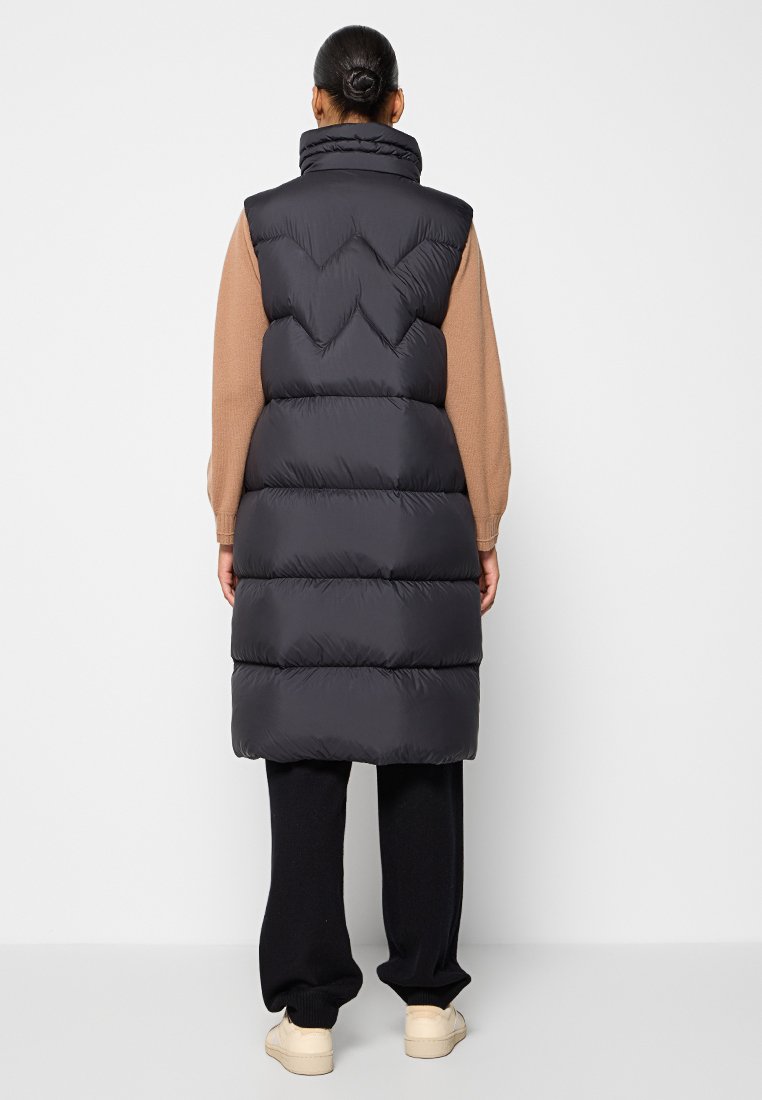 Puffer-Weste in Schwarz ohne Ärmel mit hohem Kragen, ausgestattet mit horizontalen Steppnähten und einer glatten, glänzenden Textur, getragen über einem beigen Pullover.