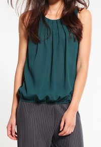 Blouse sans manches bleu sarcelle en tissu léger, avec des plis au niveau de l'encolure et une taille froncée, associée à un pantalon gris à fines rayures.