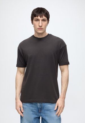 Ung mand med kort mørkt hår iført en enkel sort t-shirt og blå jeans, står foran en lysegrå baggrund.