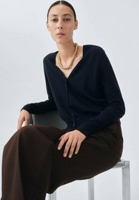 Maglione navy con scollo a V, maniche lunghe e bottoni frontali; abbinato a pantaloni larghi marroni, seduto su una sedia argentata.