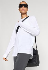 Λευκό πουκάμισο με μακριά μανίκια, γκρι ριγωτοί κολάν και μαύρη τσάντα ώμου. Μοντέλο με υπερμεγέθη μαύρα γυαλιά ηλίου, μαλακό ύφασμα, casual εφαρμογή.