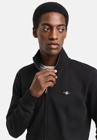 Sudadera negra con cremallera y cuello alto, con un pequeño logo bordado en el pecho y puños y bajo acanalados. Textura suave.