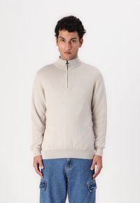 Beige Reißverschluss-Pullover mit hohem Kragen sowie gerippten Bündchen und Saum, aus glattem Strickstoff gefertigt und mit einem dezenten Logo auf der Brust.