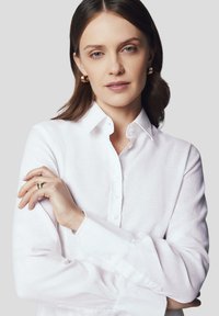 Biała koszula na guziki wykonana z lekkiego materiału, z klasycznym kołnierzykiem, długimi rękawami i luźnym krojem. Widoczna drobna tekstura.