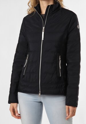 Kvinde iført en tætsiddende sort quiltet jakke med sølvfarvede lynlåse og lyseblå jeans, stående mod en ensfarvet baggrund.