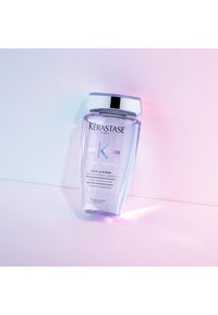 Bouteille de shampoing violet Kérastase Blond Absolu Bain Lumière avec bouchon argenté sur une surface rose pastel et bleue, inclinée vers la droite.