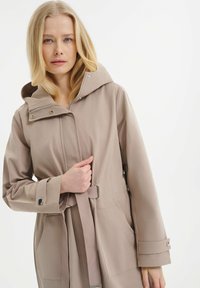 Helle beige Regenjacke mit Kapuze, tailliert geschnitten, zwei seitlichen Taschen und Knopfakzenten an den Bündchen. Glatte Stofftextur.