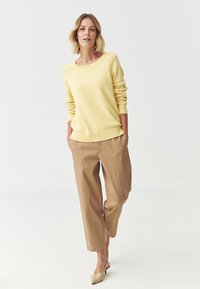 Pulover tricotat de culoare galben deschis, cu manșete și tiv cu dungi, asortat cu pantaloni bej, largi. Modelul poartă pantofi cu toc mic, bej.
