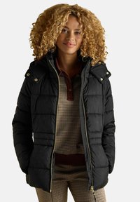 Kourtney blonde juustega naine, kes kannab musta tikkjakki pruunikate ruuduliste särkide ja pükste kohal, neutraalse tausta ees.