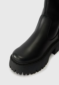 Crna kožna gležnjača sa zaobljenim prstom, masivnim rebrastim đonom i bočnom elastičnom pločom. Glatka tekstura i dizajn viskog kontrasta.