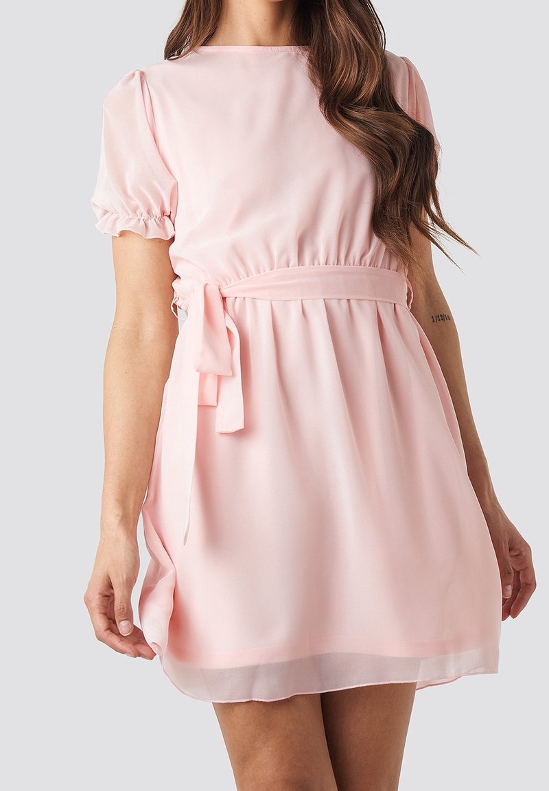 Femme portant une robe rose clair à manches courtes avec une ceinture à la taille et des bords de manches volantés sur un fond gris uni.