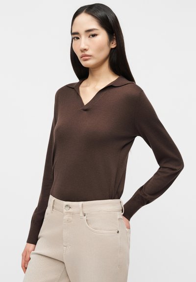 Femme aux longs cheveux noirs portant un haut marron ajusté à manches longues et un pantalon taille haute beige clair, debout devant un fond uni.