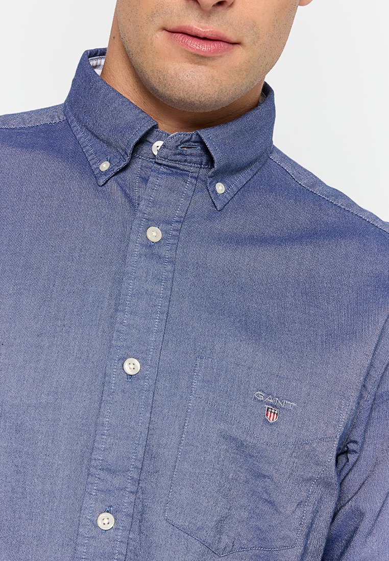 Camisa azul de botón con tejido texturizado; presenta un cuello blanco, un bolsillo frontal con logo y una tapeta de botones estándar con botones redondos.