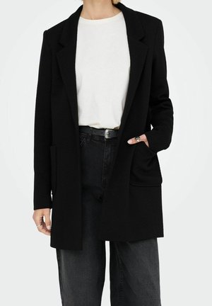 Persoon die een zwarte lange blazer draagt, een wit t-shirt, zwarte broek en een decoratieve riem met één hand in het zakje van de blazer.