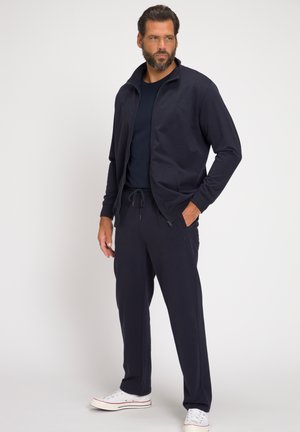 Marinefarbener Trainingsanzug mit Reißverschlussjacke und Jogginghose mit Kordelzug, aus glattem Stoff gefertigt. Verfügt über Taschen und ein dezentes Logo. Weiße Schuhe runden das Outfit ab.