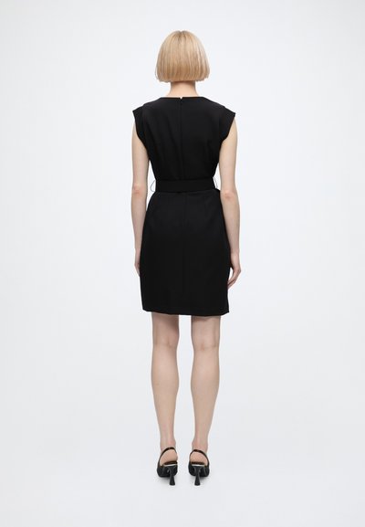 Femme aux cheveux blonds courts portant une robe noire sans manches et des sandales noires à talons, debout de dos sur un fond blanc uni.