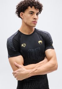 Νεαρός με σγουρά μαλλιά, φοράει μαύρο αθλητικό πουκάμισο με χρυσούς δράκους και λογότυπα με χαυλιόδοντες, με σταυρωμένα τα χέρια, κοιτάζοντας προς τα πλάγια.