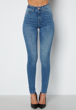 Jean bleu taille haute en denim extensible, avec une silouhette ajustée et un design épuré, associés à des talons hauts noirs.