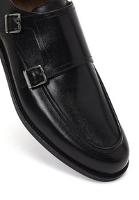 Zapato de doble hebilla de cuero negro con acabado brillante, hebillas plateadas, punta redonda y detalles cosidos a lo largo de la suela.