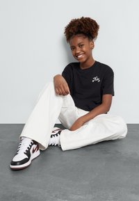 Černé tričko Nike SB, volné bílé kalhoty a černo-červené tenisky. Osoba sedí, usmívá se, má kudrnaté vlasy stylizované do drdolu. Šedé pozadí.