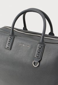 Sac en cuir gris avec une finition texturée, équipé de poignées doubles et de quincaillerie en tonalité argent. Logo "TOMMY HILFIGER" affiché sur le côté.