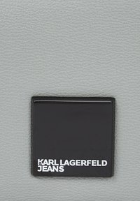 Juodas stačiakampis žymeklis su iškiliais baltais užrašais "KARL LAGERFELD JEANS" ant tekstūrinio pilkos odos fono.