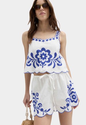 Vrouw draagt een wit mouwloos crop top en shorts met blauwe bloemenborduursels, houdt een gevlochten tas vast en draagt een zonnebril.