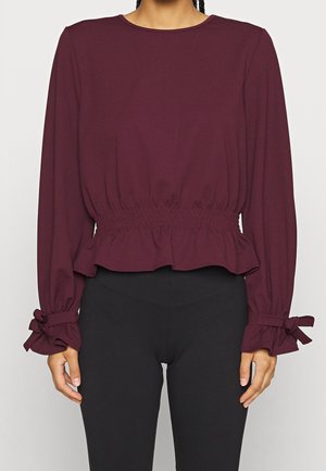 Kobieta ubrana w bordową bluzkę z długim rękawem, z taliowanym stanem i falbanianymi mankietami związanymi kokardkami, w połączeniu z czarnymi spodniami.