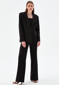 Svart skräddarsydd kostym med en kavaj med sned krage, enkel knappstängning och vida byxor, kompletterad med ett silversmycke.