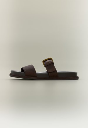 Σκούρο καφέ δερμάτινο σανδάλι με δύο λουριά, ένα φαρδύ πάνω από τα δάχτυλα και ένα με μεταλλική πόρπη πάνω από την καμάρα του ποδιού, σχεδιασμένο με επίπεδη σόλα.
