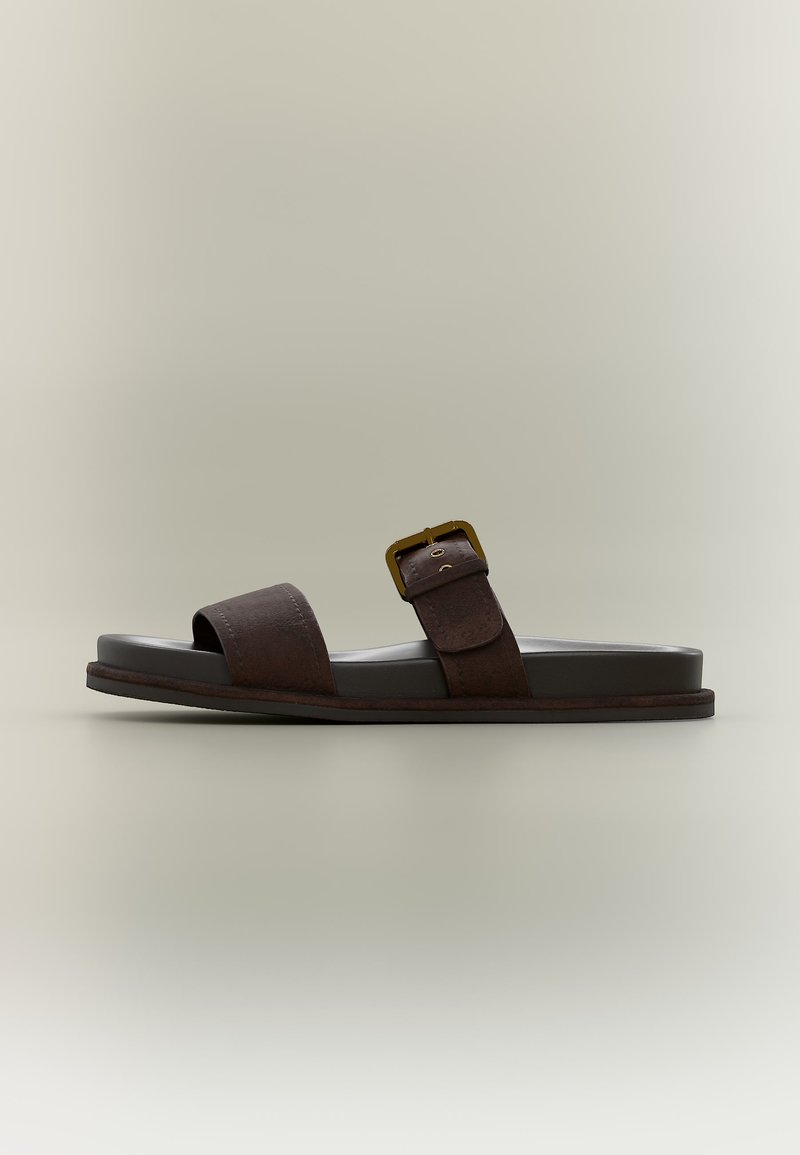 Σκούρο καφέ δερμάτινο σανδάλι με δύο λουριά, ένα φαρδύ πάνω από τα δάχτυλα και ένα με μεταλλική πόρπη πάνω από την καμάρα του ποδιού, σχεδιασμένο με επίπεδη σόλα.
