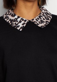 Persona que lleva una blusa negra con un cuello puntiagudo que presenta un patrón de leopardo en tonos beige y negro.
