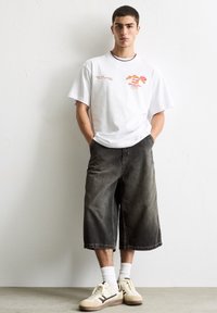 Hvid kortærmet t-shirt med farverig tekst, parret med løse, mørke denimshorts til knæene og beige sneakers med brune accenter.