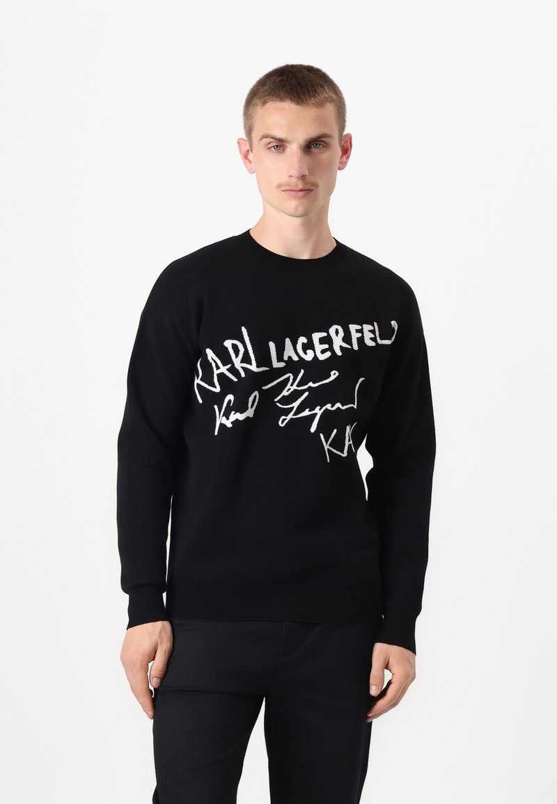 Black sweater with white text featuring signatures and branding. Crew neck, long sleeves, ribbed cuffs, and hem. Soft knit texture.