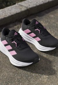 Czarne i różowe buty sportowe z grubymi białymi podeszwami spoczywające na wybrukowanej nawierzchni na zewnątrz, blisko betonowego krawężnika.