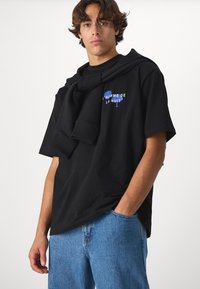 Svart t-shirt med kort ärm med blå text "L'HOMME DE LA NUIT" på bröstet; ihop med ljusblå jeans och en svart tröja över axlarna.