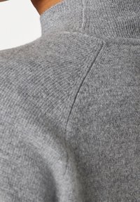 Gros plan sur l'épaule et le cou d'une personne portant un pull en maille texturée gris clair avec des détails de couture visibles.