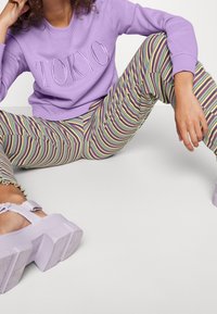 Persona seduta con le gambe divaricate, che indossa una felpa lavanda con la scritta "TOKYO", pantaloni a righe multicolori e sandali chunky lavanda.
