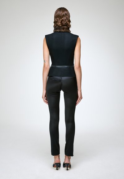 Femme debout, de dos, portant un haut noir sans manches avec un col en fourrure, un pantalon noir ajusté et des chaussures à talons noires sur un fond uni.