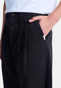 Schwarze Baumwollhosen mit einer strukturierten Passform, versehen mit vorderen Falten, seitlichen Taschen und einem kleinen weißen Etikett in der Nähe der Tascheneröffnung.