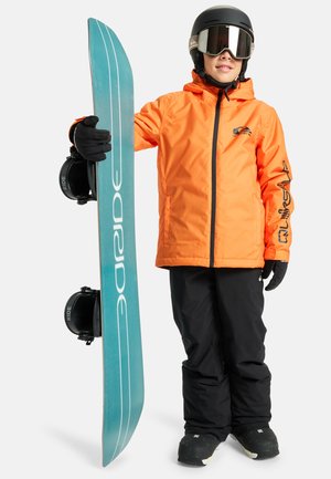 Niño con casco negro, gafas, chaqueta naranja y pantalones negros sostiene una tabla de snowboard azul en posición vertical sobre un fondo blanco.