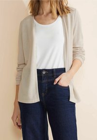 Ljust beige kofta med lös passform, öppen framdel och ribbad struktur, kombinerad med en vit topp och mörkblå högmidjade jeans.