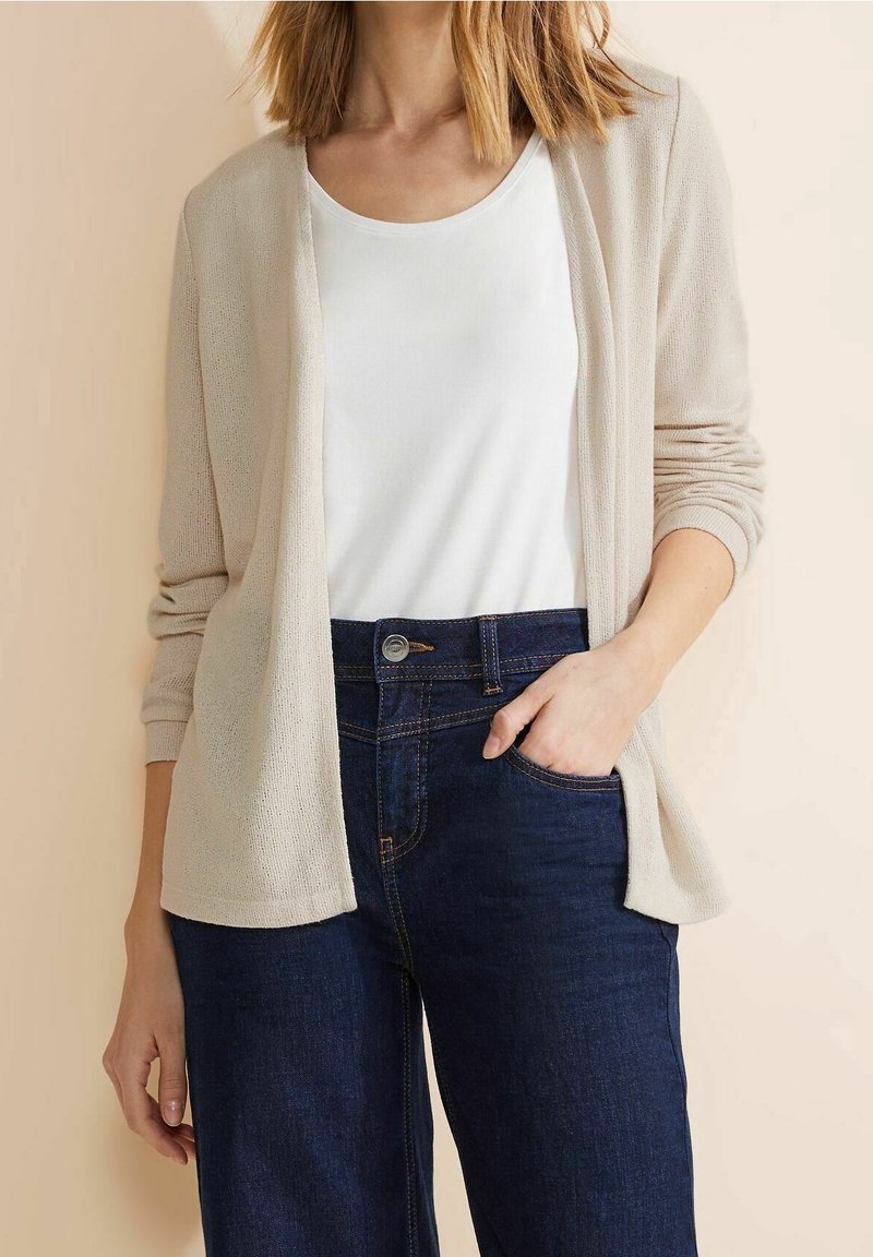 Ljust beige kofta med lös passform, öppen framdel och ribbad struktur, kombinerad med en vit topp och mörkblå högmidjade jeans.