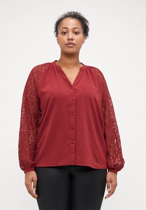 Blouse rouge avec des manches longues en dentelle, devant à boutons et encolure ronde. Tissu lisse avec un motif texturé. Pantalons noirs visibles en bas.