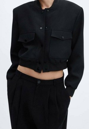 Chaqueta negra recortada con botones de presión frontales y dos bolsillos con solapa, combinada con pantalones plisados negros de talle alto que dejan el abdomen al descubierto.