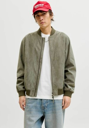 Jeune homme portant une casquette rouge, une veste bomber vert olive, un t-shirt blanc et un jean bleu clair, debout devant un fond uni clair.