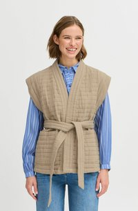 Gilet beige matelassé avec des manches larges et une ceinture à nouer, associé à une chemise à rayures bleues. Le tissu a une finition texturée et une apparence douce.