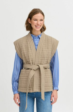 Gilet beige matelassé avec des manches larges et une ceinture à nouer, associé à une chemise à rayures bleues. Le tissu a une finition texturée et une apparence douce.