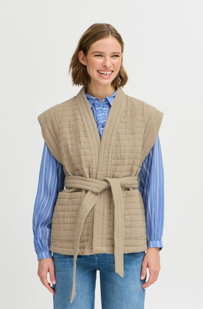 Gilet beige matelassé avec des manches larges et une ceinture à nouer, associé à une chemise à rayures bleues. Le tissu a une finition texturée et une apparence douce.